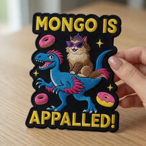 May include: Black embroidered patch with the text "MONGO IS APPALLED!" featuring a blue dinosaur with pink accents and a cat wearing sunglasses and a crown. Pink donuts and yellow stars are also included in the design.