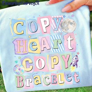 May include: A light blue t-shirt with the words "COPY HEART COPY BRACELET" in colorful, block letter style. The design includes a moon, stars, a rocket ship, and a planet.