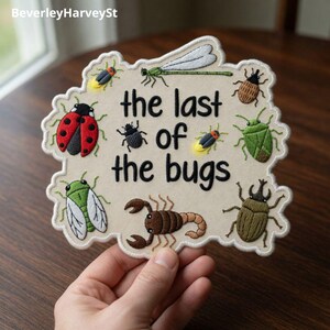 Puede incluir: Parche bordado beige con el texto "the last of the bugs" rodeado de varios insectos bordados, incluyendo una mariquita, una libélula y un escarabajo. El parche tiene un borde festoneado.