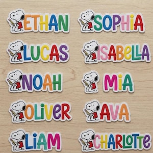 May include: Ten embroidered patches featuring the cartoon character Snoopy holding a pencil and a red book, each paired with a different name in colourful, bold, block-style lettering including Ethan, Sophia, Lucas, Isabella, Noah, Mia, Oliver, Ava, Liam, and Charlotte.