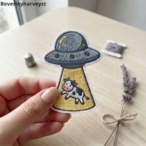 May include: An embroidered patch featuring a grey UFO with yellow stars and a yellow tractor beam pulling up a black and white cow. The patch has a white border and is shaped like a flying saucer. The text Beverleyharveyst is visible in the corner.
