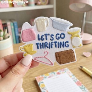 以下が含まれることがあります： LET'S GO THRIFTINGという青い文字が刺繍された白いワッペン。ピンクのバッグ、クリーム色のマグカップ、水色のボウル、黄色のティーカップ、黄色の秤、ピンクのハンガー、茶色のチェストといった様々なアイコンが周囲に配置されています。
