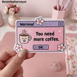 May include: A rectangular embroidered patch featuring a light purple and pink computer pop-up window design. The text reads Warning! You need more coffee. with an OK button. It includes a smiling coffee mug graphic and three white flower accents.