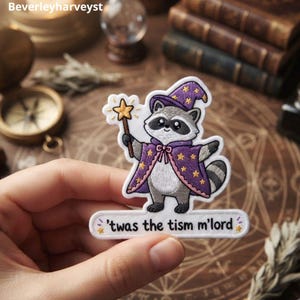 Puede incluir: Un parche bordado que muestra un mapache gris y blanco vestido de mago con una túnica morada con estrellas amarillas y un sombrero a juego, sosteniendo una varita de estrella amarilla. Debajo del mapache hay una banda blanca con el texto 'twas the tism m'lord'.