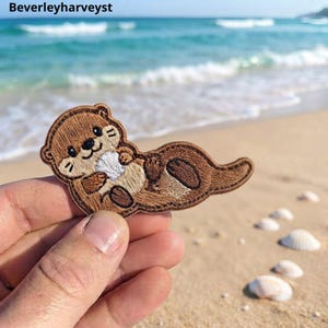 Può includere: Una toppa ricamata raffigurante una lontra marrone con il ventre color crema, che tiene in mano una piccola conchiglia bianca. La lontra presenta dettagli ricamati in marrone scuro per occhi, naso e zampe, con un contorno netto. Il testo Beverleyharveyst è visibile.