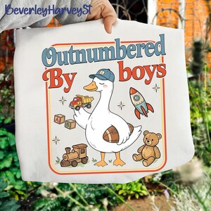 May include: White tote bag with a cartoon goose wearing a baseball cap and holding a toy truck. The bag features the text "Outnumbered By boys" in a retro font, along with illustrations of a rocket, teddy bear, and train.