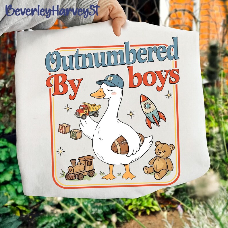 May include: White tote bag with a cartoon goose wearing a baseball cap and holding a toy truck. The bag features the text "Outnumbered By boys" in a retro font, along with illustrations of a rocket, teddy bear, and train.