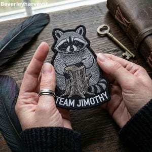 May include: An embroidered patch featuring a grey and black raccoon sitting behind a brown tree stump. The raccoon has a striped tail and a black mask around its eyes. Below the raccoon, the text "TEAM JIMOTHY" is stitched in white letters.