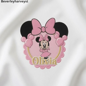May include: An embroidered patch on white fabric featuring a circular design with a scalloped pink border. The center shows a cartoon mouse character in a pink dress and bow, surrounded by pink balloons, a large pink bow, and the name Olivia.