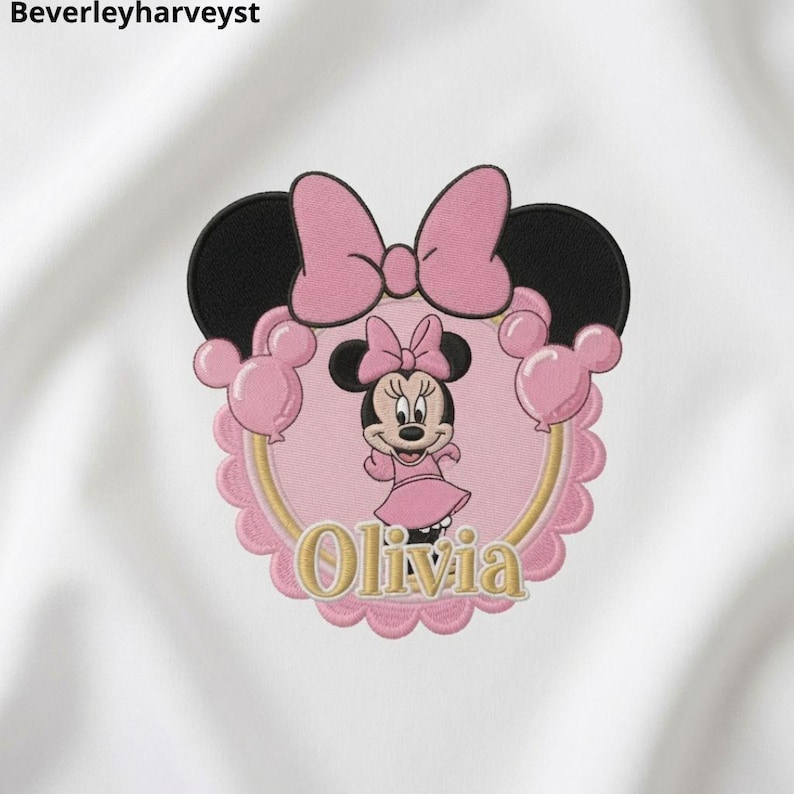May include: An embroidered patch on white fabric featuring a circular design with a scalloped pink border. The center shows a cartoon mouse character in a pink dress and bow, surrounded by pink balloons, a large pink bow, and the name Olivia.
