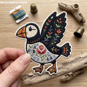 Puede incluir: Un parche bordado de un frailecillo con el pecho color crema, una rosa rosa central y detalles florales azules. Las alas y la espalda son negras con bordados coloridos de hojas y flores. El ave tiene un pico naranja y negro y patas naranjas.