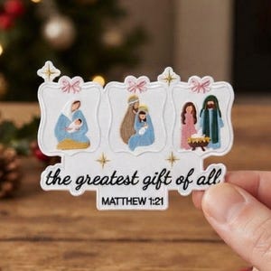 Può includere: Un adesivo bianco fustellato con tre pannelli rettangolari che mostrano illustrazioni in stile ricamo della Natività, sormontati da fiocchi rosa e stelle dorate. Il testo recita "the greatest gift of all" e "MATTHEW 1:21" in carattere nero.