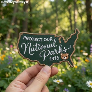 Op de afbeelding: Een donkergroene geborduurde patch in de vorm van de kaart van de Verenigde Staten met een beige rand. Het bevat de witte tekst "PROTECT OUR National Parks" en "1916" naast een klein, bruin geborduurd berengezichtje rechtsonder.