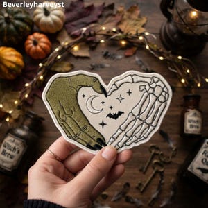 Könnte beinhalten: Ein herzförmiger Stickaufnäher mit einer grünen Monsterhand auf der linken und einer weißen Skeletthand auf der rechten Seite, die zusammen ein Herz bilden. In der Mitte befinden sich ein weißer Halbmond, eine kleine schwarze Fledermaus und Sterne.