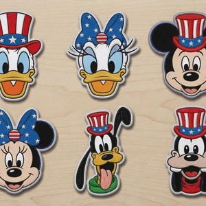 Könnte beinhalten: Ein Set aus sechs bestickten Aufnähern mit Zeichentrickfiguren-Gesichtern. Die Designs zeigen Donald Duck, Daisy Duck, Mickey Maus, Minnie Maus, Goofy und Pluto, alle verziert mit patriotischen rot-weiß-blauen Sternen- und Streifenmotiven.