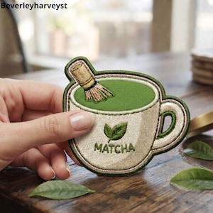 May include: An embroidered patch shaped like a cup of matcha tea. The cup is cream-colored with a green interior, featuring a brown bamboo whisk and a green leaf icon above the text MATCHA. The patch has a pink border outline.