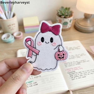 Puede incluir: Un parche bordado que muestra un fantasma blanco con un lazo rosa oscuro en la cabeza, mejillas rosadas y una cara sonriente. El fantasma sostiene una pequeña calabaza rosa en una mano y un lazo rosa de concienciación en la otra, con borde negro.
