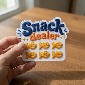 May include: A white embroidered patch featuring the dark blue text Snack and orange text dealer. Below the text are six small yellow fish-shaped crackers arranged in two rows of three, accented by small yellow star and dash embroidery details.