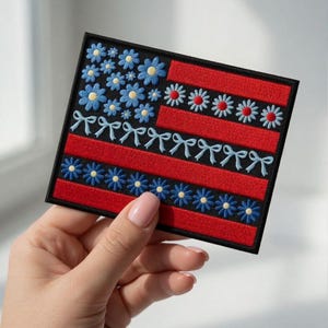 Può includere: Toppa ricamata con un design stilizzato della bandiera americana. La bandiera incorpora strisce rosse, un campo nero con dettagli floreali blu e file di fiocchi bianchi. La toppa ha un bordo nero e il testo "BEVERLEYHARVEYST" in alto.