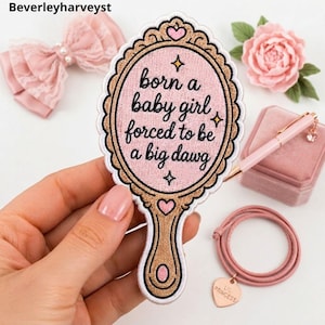 May include: An embroidered patch shaped like a hand mirror with a gold frame and a light pink center. The text reads, "born a baby girl forced to be a big dawg," with small star accents and two pink hearts. The patch features a scalloped edge.
