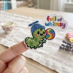 May include: An embroidered patch featuring a green caterpillar wearing a blue cap and holding a colorful rainbow lollipop. A white speech bubble above the caterpillar contains the multi-colored embroidered text full of whimsy.