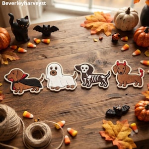 May include: Four embroidered dachshund patches featuring Halloween costumes: a brown dog in a vampire cape, a dog under a white ghost sheet, a dog with a skeleton print, and a brown dog with red devil horns and a tail, all arranged in a row.