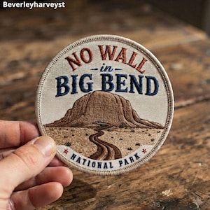 Puede incluir: Un parche bordado circular con fondo crema que muestra un paisaje montañoso marrón y un camino sinuoso. El texto NO WALL in BIG BEND NATIONAL PARK está bordado en hilo azul oscuro y color óxido con dos pequeñas estrellas rojas.
