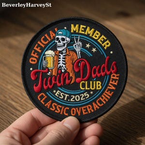 May include: Embroidered patch with a black background featuring a skeleton wearing a cap and orange jacket, holding a beer. Text reads "OFFICIAL MEMBER Twin Dads CLUB CLASSIC OVERACHIEVER EST. 2025".