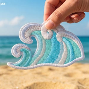 Könnte beinhalten: Gestickter Wellenaufnäher in Weiß, Hellblau und Türkis. Der Aufnäher wird vor dem Hintergrund eines Strandes und Ozeans gehalten. Der Aufnäher hat einen weißen Rand und ist ein dekorativer Artikel.