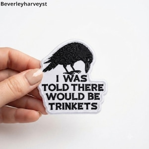 Könnte beinhalten: Ein weißer gestickter Aufnäher mit einer schwarzen Rabenillustration über dem schwarzen Text I WAS TOLD THERE WOULD BE TRINKETS. Der Aufnäher hat eine gestanzte Form, die der Kontur des Vogels und des Textblocks folgt.