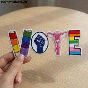 May include: An embroidered patch spelling VOTE. The V is a stack of books in red, orange, yellow, and white. The O is a blue raised fist in a circle. The T is a pink uterus with a heart. The E features horizontal stripes of rainbow colors.