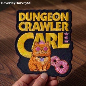 Puede incluir: Parche bordado con el texto "DUNGEON CRAWLER CARL" en amarillo, encima de un gato de dibujos animados con gafas de sol y una corona. El gato es naranja con una pajarita morada, y hay donuts rosas en la parte inferior derecha.