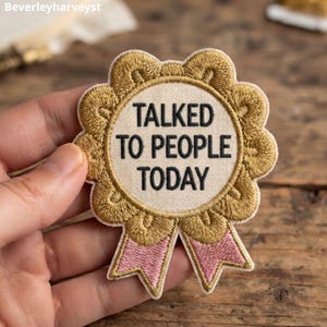 Puede incluir: Un parche bordado circular con forma de cinta de premio con un centro color crema que presenta el texto negro TALKED TO PEOPLE TODAY. El borde es de hilo dorado con un borde festoneado y dos colas de cinta rosa en la parte inferior.