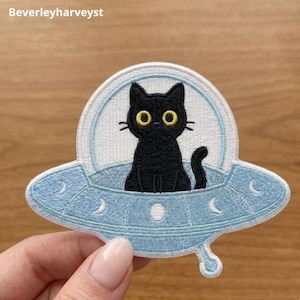 May include: An embroidered patch featuring a black cat with large yellow eyes sitting inside a light blue UFO. The spacecraft has a white dome, a circular base with crescent moon and circle details, and the text Beverleyharveyst at the top.