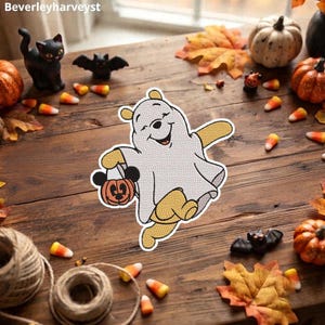 May include: An embroidered patch featuring a cartoon bear dressed as a ghost, wearing a white sheet with eye holes, holding a small orange pumpkin bucket with black mouse ears. The bear has yellow limbs and a smiling face, with a white border.