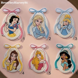 May include: Embroidered patches featuring Disney princesses Snow White, Elsa, Cinderella, Belle, Aurora, and Jasmine. Each patch has a portrait of a princess, a ribbon, and a pearl-like border. The patches are on a pink surface.