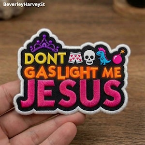 Op de afbeelding: Een geborduurde patch met een zwarte achtergrond en witte rand met de tekst DONT GASLIGHT ME JESUS in geel, oranje en roze. Boven de tekst staan kleine iconen van een paarse kroon, een witte boxershort met hartjes, een schedel, een blauwe dinosaurus en een roze bom.