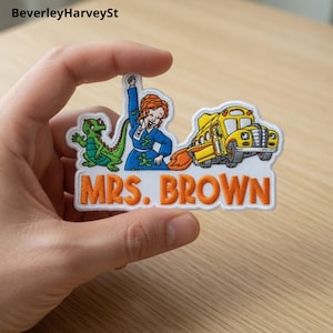 Könnte beinhalten: Gestickter Aufnäher mit einem Cartoon-Design einer Frau in einem blauen Kleid, einem grünen Dinosaurier und einem gelben Schulbus mit Flammen. Der Text "MRS. BROWN" steht in orangefarbenen Blockbuchstaben.