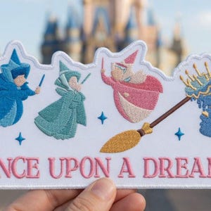 Può includere: Toppa ricamata con tre personaggi delle fate in blu, verde e rosa, con una scopa d'oro e la scritta "ONCE UPON A DREAM" in rosa. La toppa è bianca con un castello blu sullo sfondo.