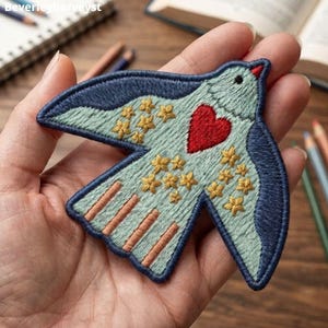 May include: An embroidered bird patch featuring a light blue body, dark blue wings, and a red heart on its chest. The design includes small yellow star-shaped accents across the wings and body, with light orange vertical stripes on the tail.