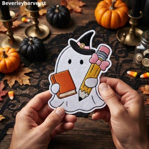 Op de afbeelding: Een geborduurde patch met een witte geest die een zwarte heksenhoed met een roze band draagt. De geest houdt een oranje boek in de ene hand en een geel potlood met een roze gum en strik in de andere hand. Het ontwerp heeft een witte rand.