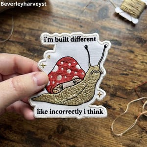 May include: An embroidered patch on white fabric featuring a tan snail with a red and white spotted mushroom shell. The text reads i'm built different above the snail and like incorrectly i think below it, accented by small gold embroidered stars.