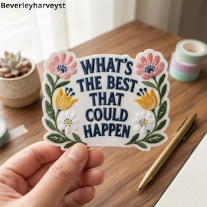 May include: A white embroidered patch with a scalloped edge featuring the dark blue text WHAT'S THE BEST THAT COULD HAPPEN. The design is framed by colourful floral motifs including pink, yellow, and white flowers with green leaves and stems.