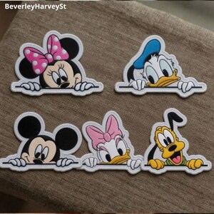 May include: Five embroidered iron-on patches featuring cartoon characters: Minnie Mouse with a pink polka-dot bow, Donald Duck in a blue cap, Mickey Mouse, Daisy Duck with a pink bow, and Pluto. Each character is shown peeking over a straight edge.