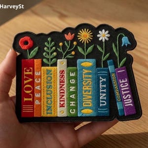 Puede incluir: Parche bordado con una fila de libros coloridos con detalles florales. Cada libro muestra una palabra: Amor, Paz, Inclusión, Bondad, Cambio, Diversidad, Unidad, Compasión y Justicia. El parche tiene un borde negro y se sostiene en una mano.
