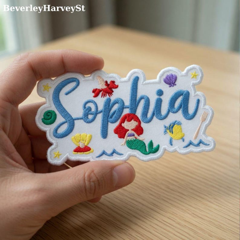 Pode incluir: Patch bordado com o nome "Sophia" em escrita azul, apresentando uma sereia, criaturas marinhas e ondas. O patch &eacute; branco com detalhes em azul, vermelho, amarelo e verde. O patch tem aproximadamente 10 cm de largura e 5 cm de altura.
