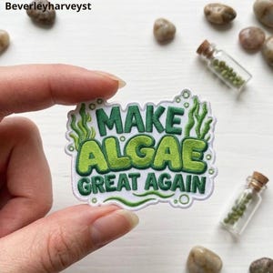 May include: An embroidered patch with a white background featuring the green text "MAKE ALGAE GREAT AGAIN" in a bold, rounded font. The design includes green seaweed illustrations and small bubbles surrounding the lettering, finished with a white border.