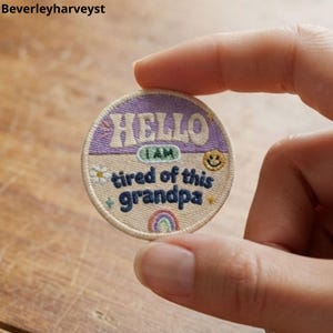 Op de afbeelding: Een ronde geborduurde patch met een lichtpaars bovengedeelte met het woord HELLO in crème, en een beige ondergedeelte met de tekst I AM tired of this grandpa in donkerblauw. Het bevat een klein madeliefje, een smiley en een regenboog.