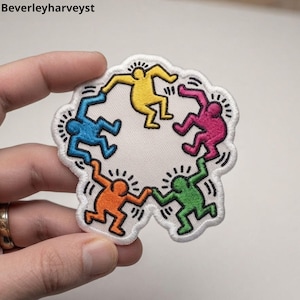 May include: An embroidered patch featuring five stylized human figures in yellow, pink, green, orange, and blue, arranged in a circle and holding hands. Each figure has black radiating lines around them, set against a white background.