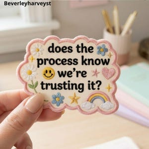 May include: An off-white embroidered patch with a scalloped pink border featuring the black text "does the process know we're trusting it?" surrounded by embroidered daisies, a yellow smiley face, a pink heart, a rainbow, and yellow stars.